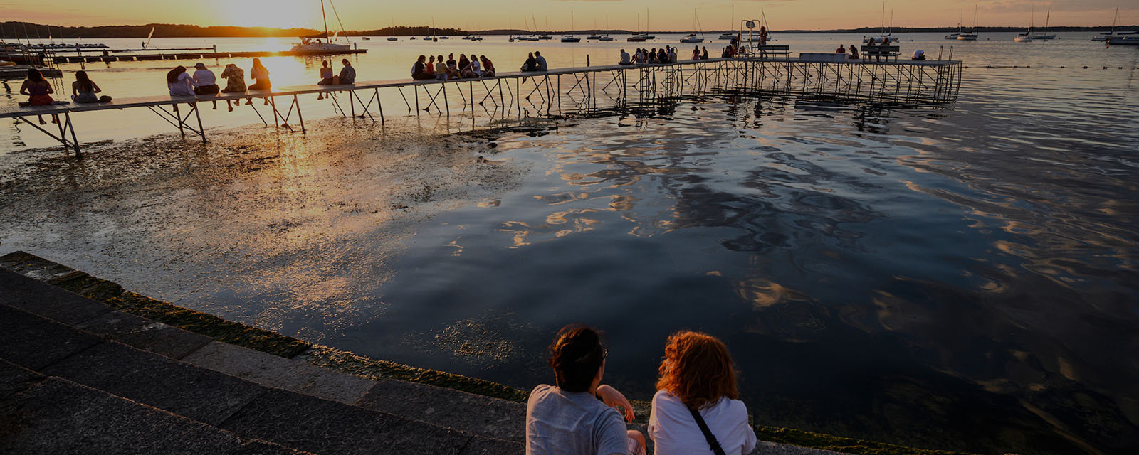 A beautiful sunset casts its reflection in the calm waters of Lake Mendota, as spectators admire it from a pier that juts out into the lake.