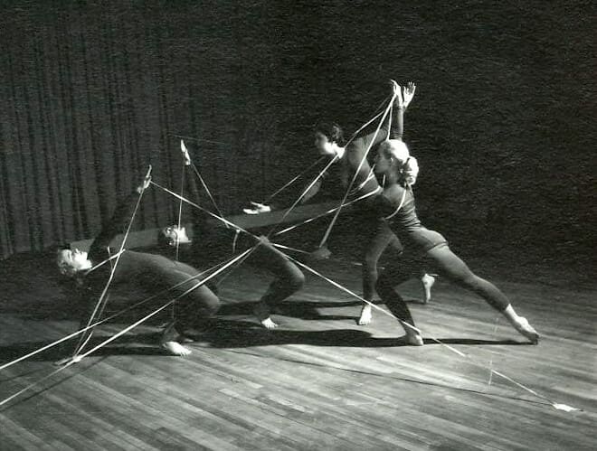 Black and white image of four female dancers, dramatically lit on stage, with ribbon wrapped around them as they engage in a dance.