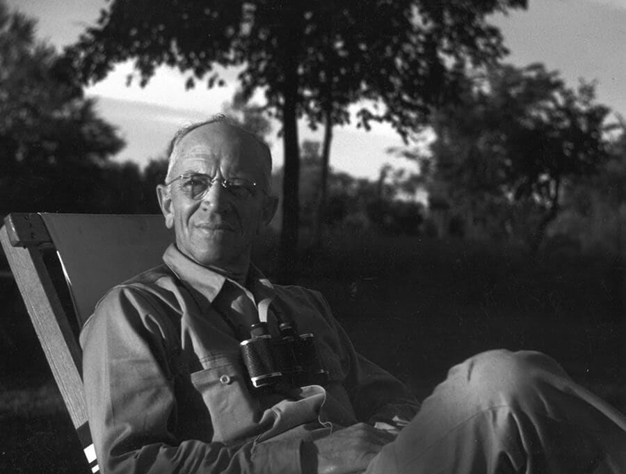 Black and white image of Aldo Leopold sitting in a canvas chair outdoors with his binoculars resting on his chest; trees in the background