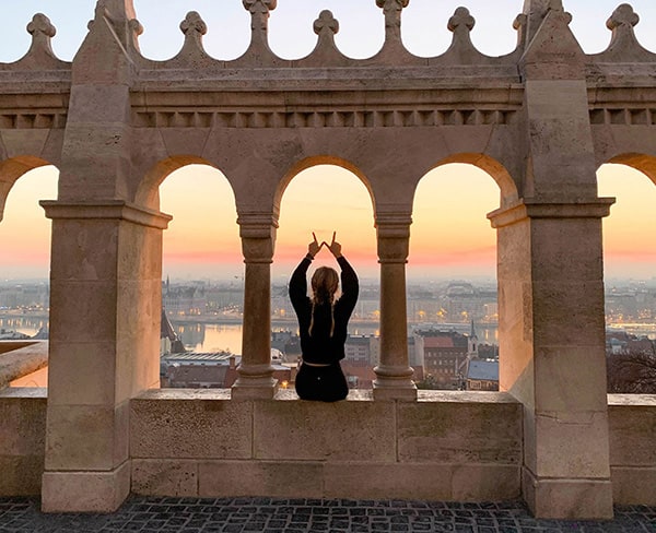 A student sits in between building columns facing a sunset with her hands above her head in the shape of the letter W