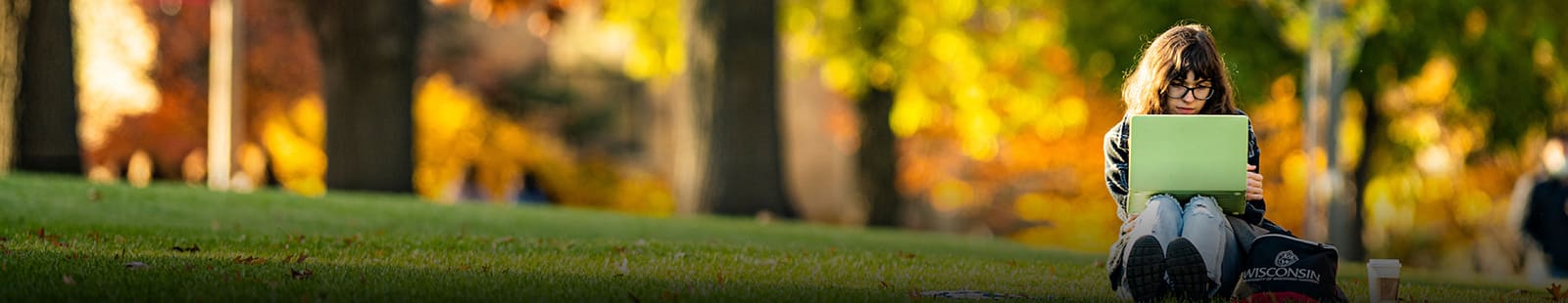 student studies with a green laptop, while sitting on the ground, a lush bokeh background of trees and greenery fills most of the scene.