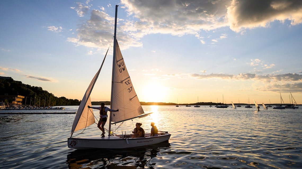 Three students are on a UW Sailing Club boat on Lake Mendota as it sails in front of the setting sun.