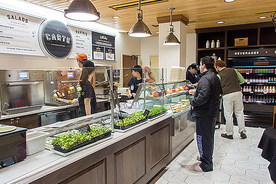 People ordering at food counter displaying greens, sandwiches and beverages with Carte logo and menu fixed to wall.