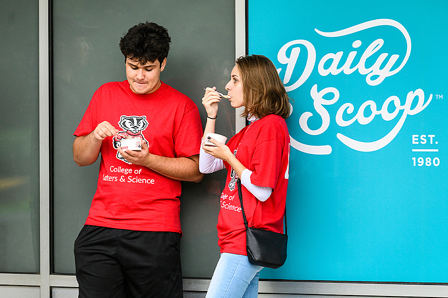 Two people in red Badger shirts eating ice cream while standing next to Daily Scoop logo, a white script text on aqua blue background.