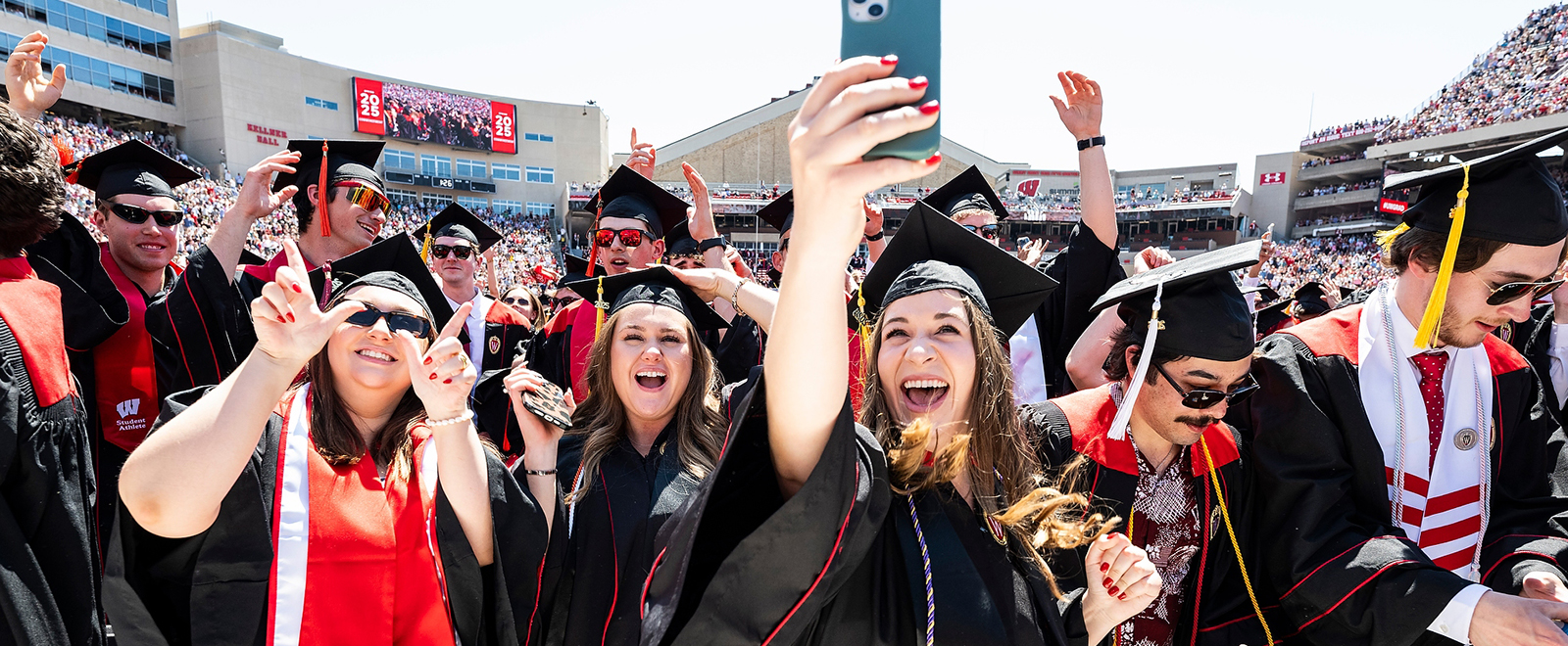 A group of graduates in black caps and gowns with red and white accents celebrate at a graduation ceremony, taking selfies and cheering in Camp Randall Stadium.