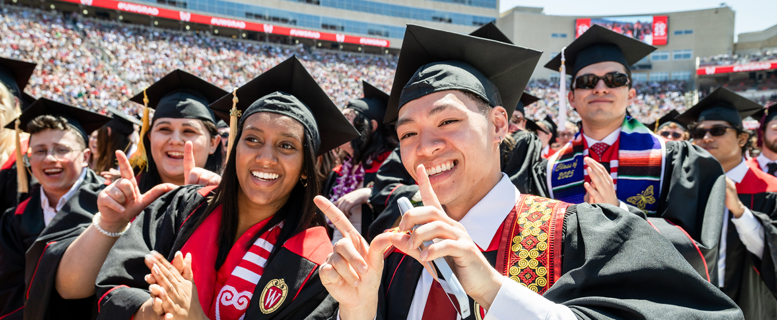 Graduates in caps and gowns smile for a photo during a commencement ceremony in a crowded stadium.