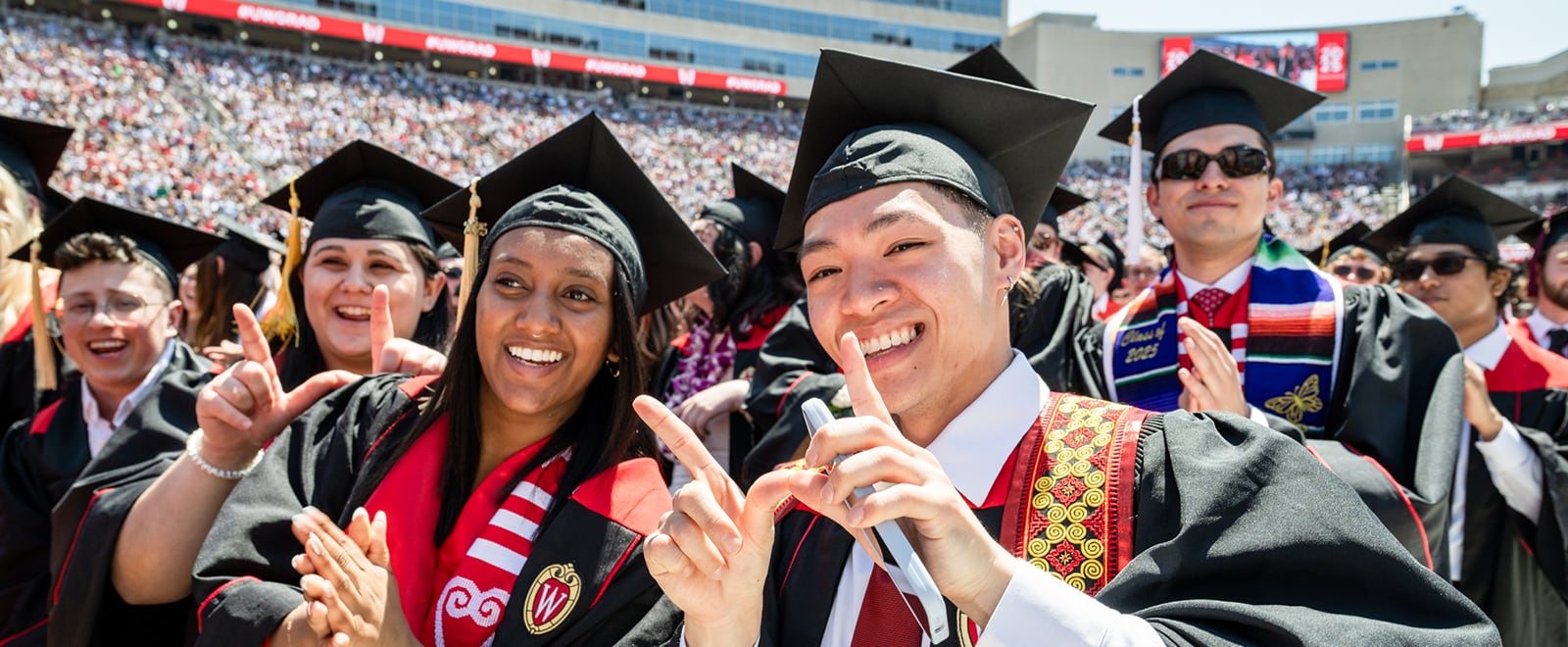 Graduates in caps and gowns smile for a photo during a commencement ceremony in a crowded stadium.