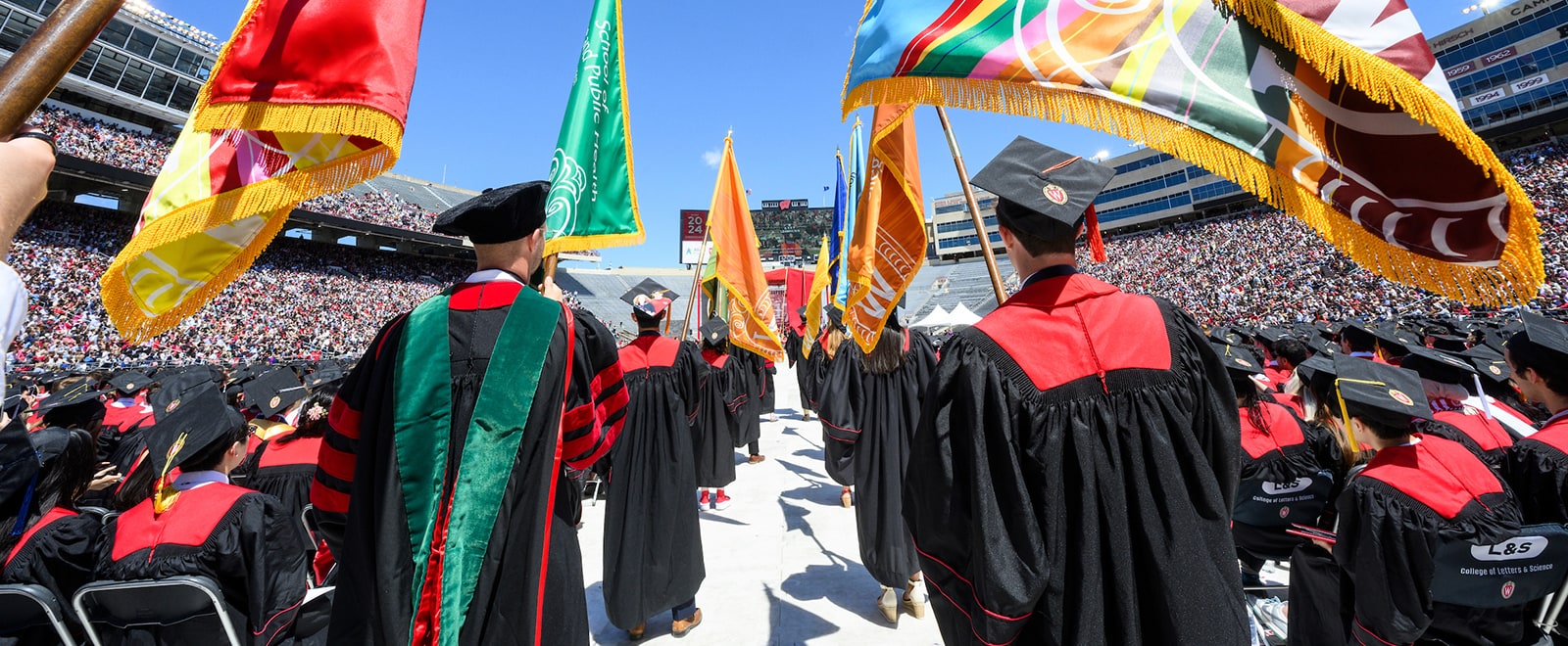 A group of graduates in caps and gowns holding colorful flags process through the center of Camp Randall. They are surrounded by seated graduates and spectators, under a clear blue sky.