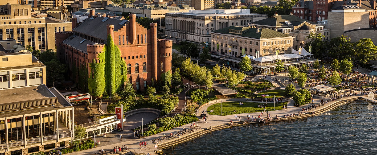 An aerial view of the lakefront near the Red Gym and Memorial Union, with lush greenery and hundreds of people enjoying the outdoor space.