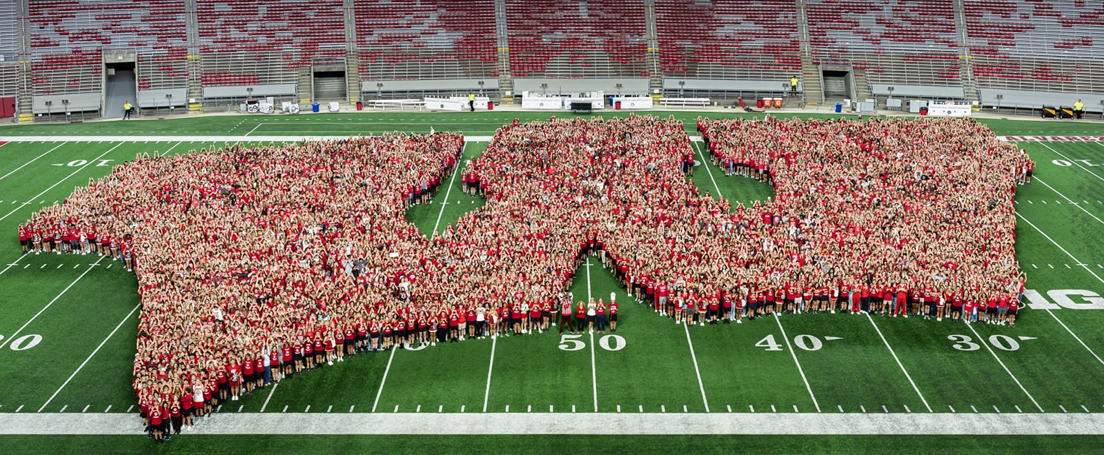 Thousands of UW students stand on the football field at Camp Randall Stadium, most wearing red t-shirts, forming the shape of UW Athletic's Motion W logo.