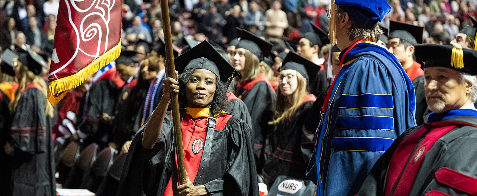 A graduate carries a flag while walking among other graduates and faculty in regalia during a commencement ceremony.