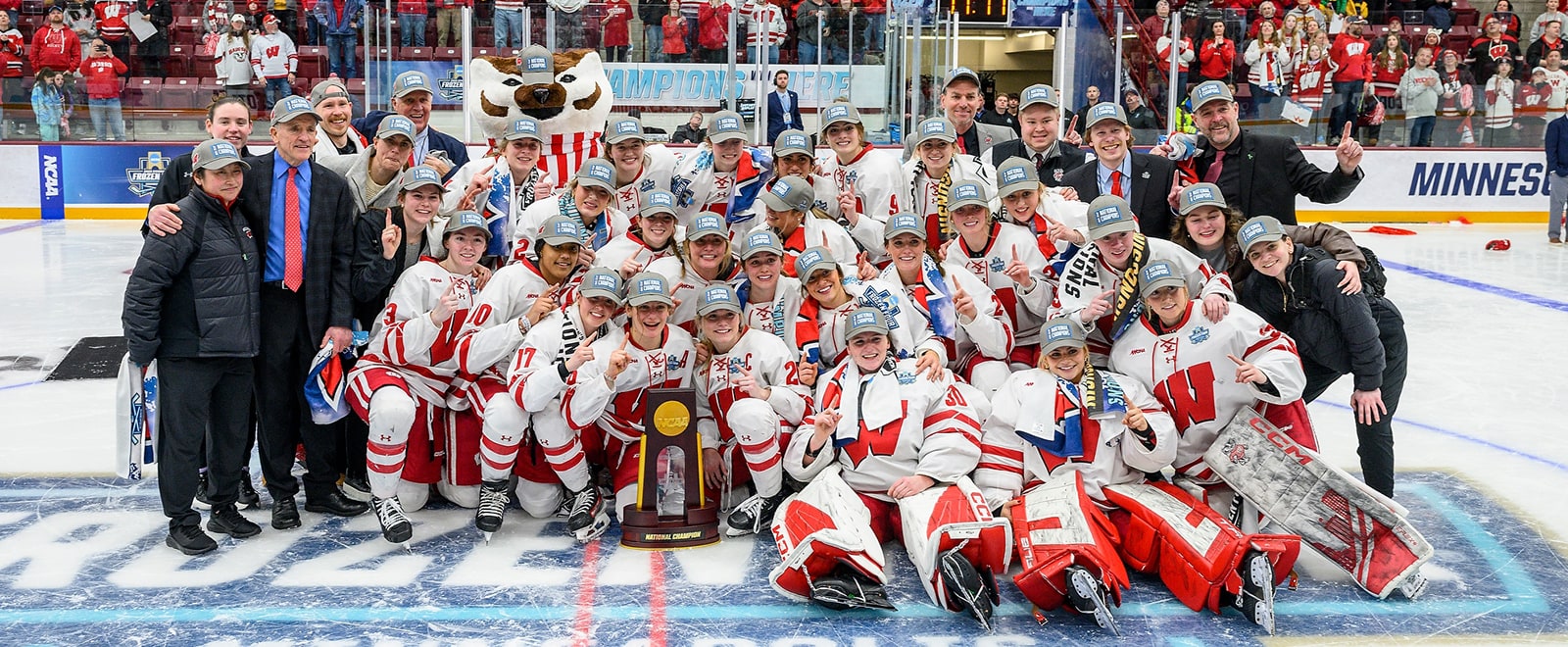 The hockey team, joined by key coaches and mascot Bucky Badger, gather in a group pose on the center of the ice. The team sits behind the NCAA national championship trophy.
