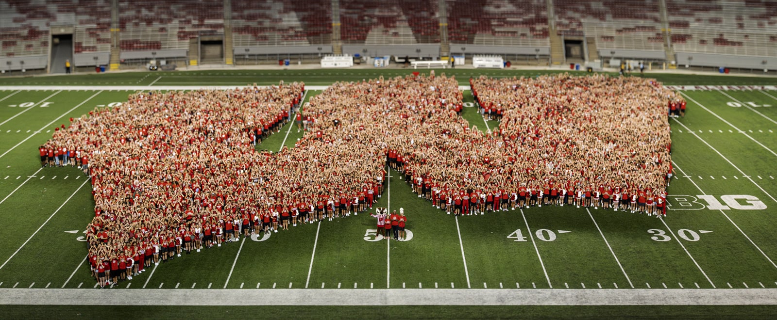 A crowd of people wearing red form a giant W on the 50 yard line in Camp Randall Stadium.
