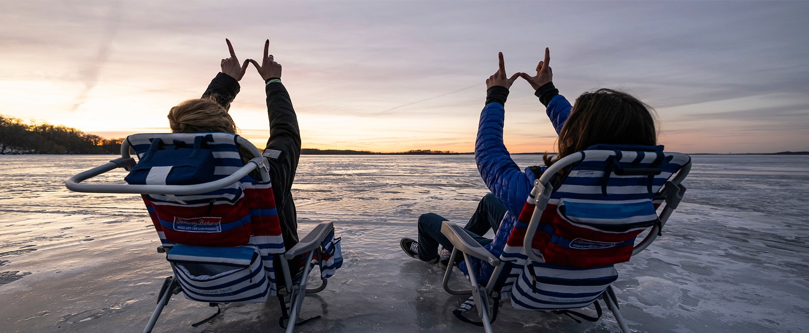Two people sit in lawn chairs on a frozen Lake Mendota. They are facing the setting sun and holding their hands above their heads in the form of the letter W.