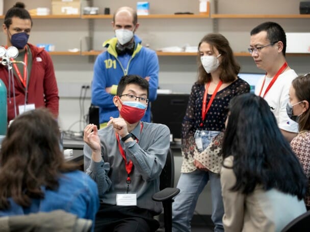 Graduate student Joseph Kim sits in the middle of a circle of scientists in a science lab, leading an interactive workshop.