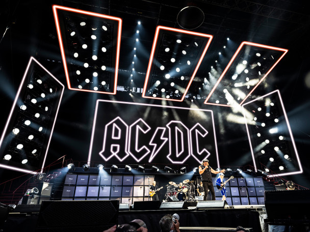 The band ACDC performs on stage in Camp Randall in front of a large neon sign that shows the band's logo.