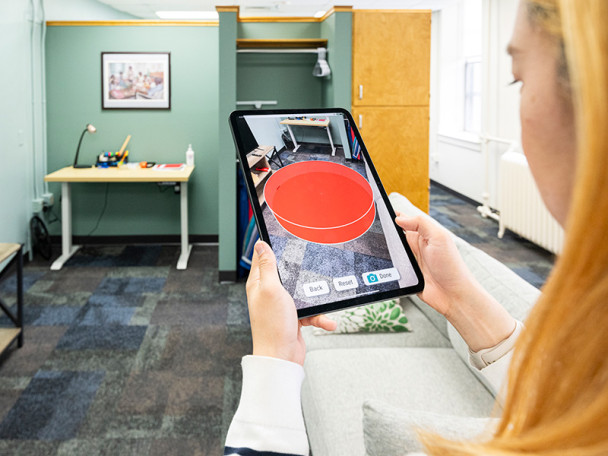 Lee holds up a iPad and sees a model of a living room through the tablet's camera. Through the view screen, Lee sees a red circle noting the area of the living room that are not accessible or may be a safety hazard to older adults.