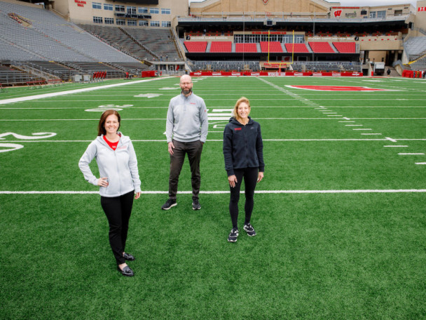 Maria Dehnert, Peter Miller, and Sara Jimenez Soffa stand at the 20 yard line in an empty Camp Randall stadium.