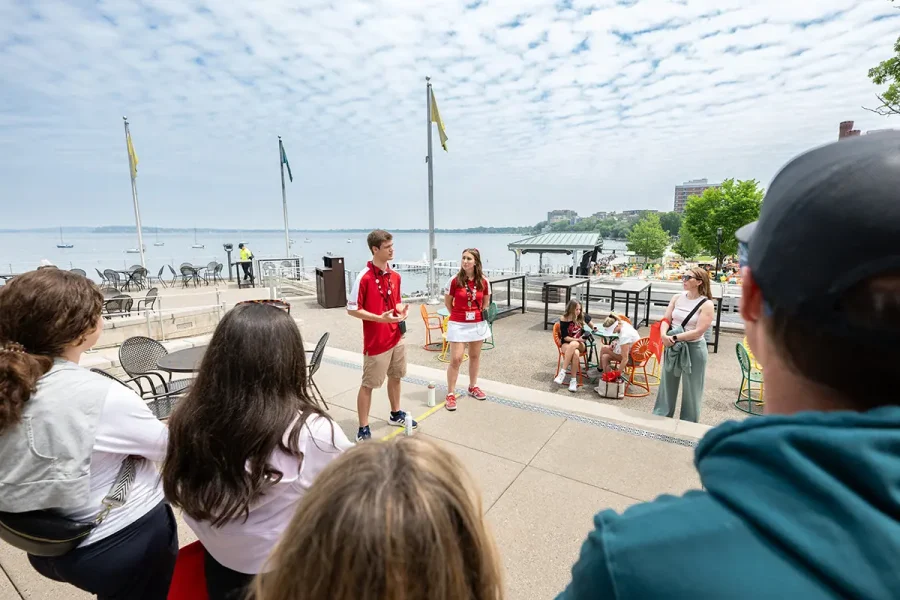Two tour guides in red shirts speak to a small group on a lakeside terrace with colorful chairs, flags, and boats visible on the water under a partly cloudy sky.