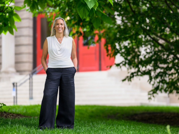 A smiling person standing with hands in their pockets in a grassy area in front of a building with a red door.
