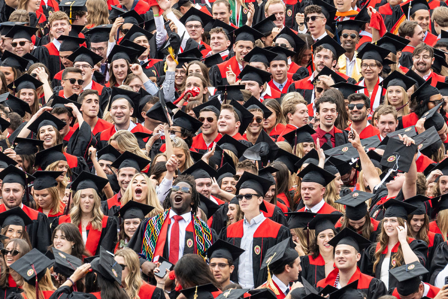 A joyful group of UW-Madison graduates in black caps and gowns with red accents celebrate together, smiling and raising their hands.