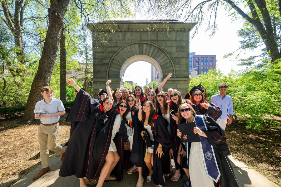 A group of people in commencement gowns pose for a picture in front of Camp Randall Arch.