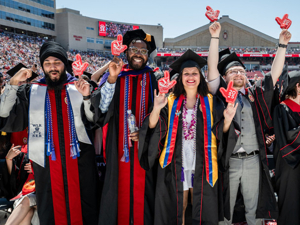 Graduates in caps and gowns celebrate in Camp Randall, holding up foam fingers that display '2025'.