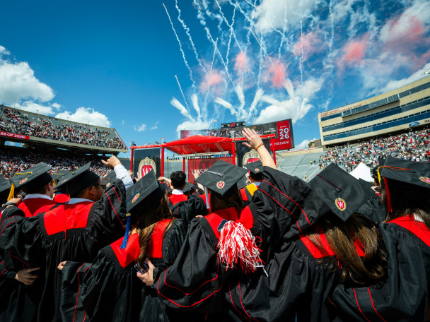Graduates in black and red caps and gowns face a stage with a digital screen reading "Spring 2026 Commencement" at a crowded outdoor stadium under a clear blue sky with white celebratory smoke trails.