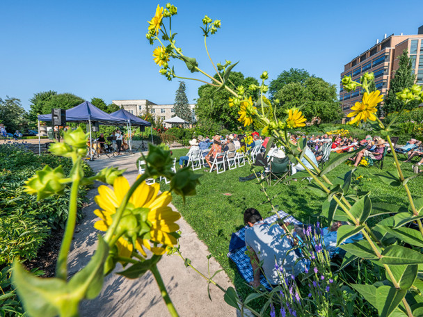 With flowers blooming in the foreground, an audience listens to a band playing in Allen Centennial Garden.