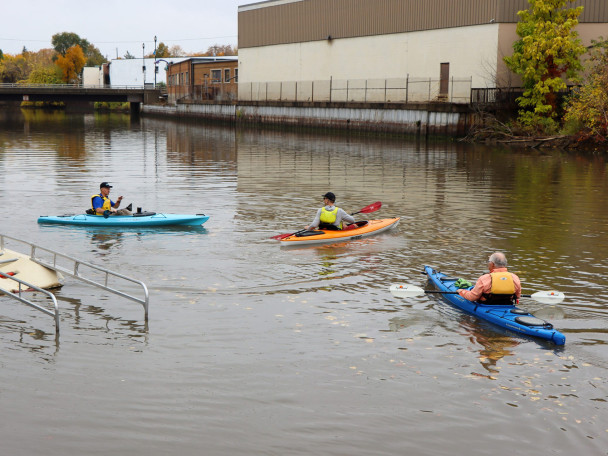 Three kayakers paddle down a calm river near an accessible dock launch, framed by autumn trees and buildings with a high-water mark visible.