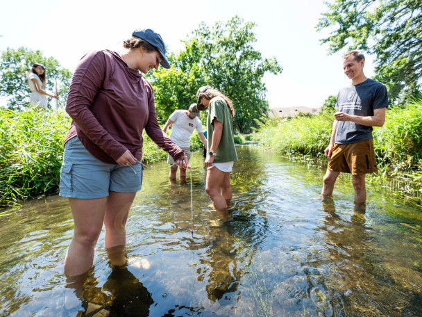 Students stand in a shallow creek. They took down at the water where they are taking samples from.