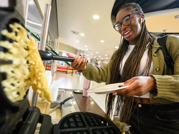 A student uses a waffle maker to prepare her breakfast. She smiles as she uses tongs to lift the waffle from the griddle.