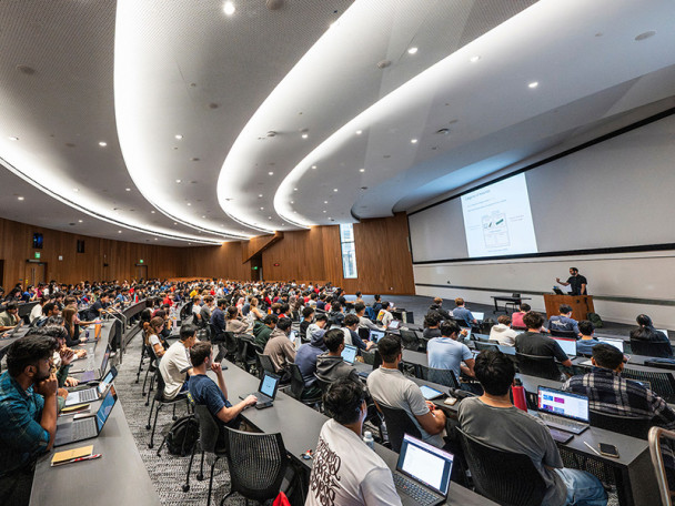 Modern classroom with many students sitting in rows with laptops facing and instructor who is lecturing behind a podium.