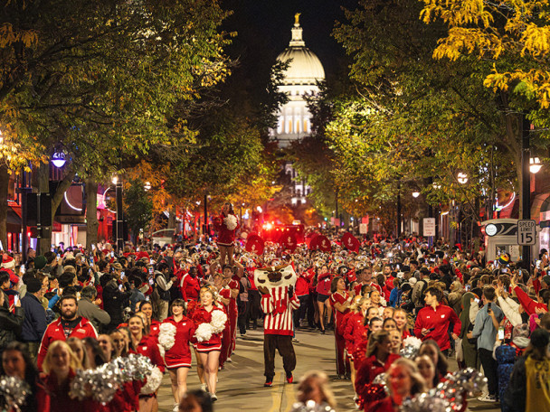 Bucky Badger leads a spirited nighttime parade down State Street, surrounded by cheerleaders and a cheering crowd, with the illuminated Wisconsin State Capitol glowing in the background.