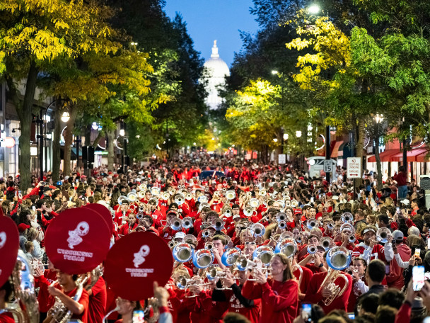 The UW Marching Band, featuring sousaphones with red covers, performs down a packed State Street during the UW Homecoming parade with the Wisconsin State Capitol in the background.