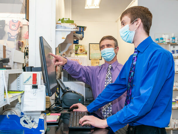 Behind the counter in a pharmacy, two men wearing button-down shirts and slacks stand in front of a computer. They are both wearing blue surgical masks. One man points to information on the computer monitor while the other types on the keyboard.