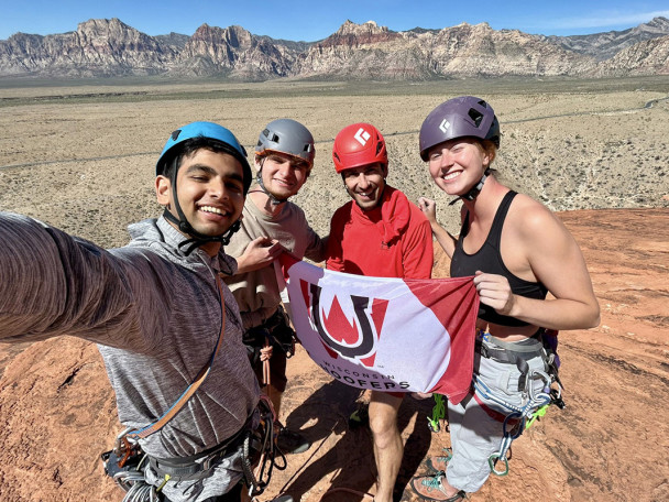 Four rock climbers wearing helmets and climbing harnesses smile for a selfie while holding a 'Wisconsin Hoofers' flag on a sandstone peak with a desert mountain range in the background.