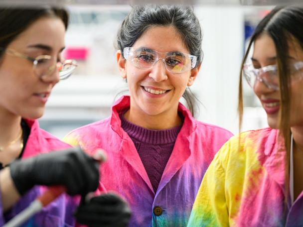 Three women in bright rainbow-colored lab coats converse in a laboratory setting.