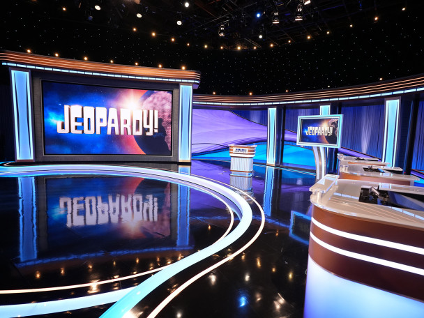 The empty studio set of the Jeopardy! game show features a large illuminated screen displaying the logo, a central host lectern, and contestant podiums under blue and purple studio lighting.