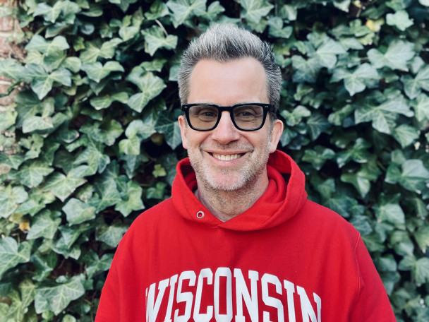 Jason Gay wears a red and white Wisconsin hoodie and smiles for a photo in front of an ivy-covered wall.