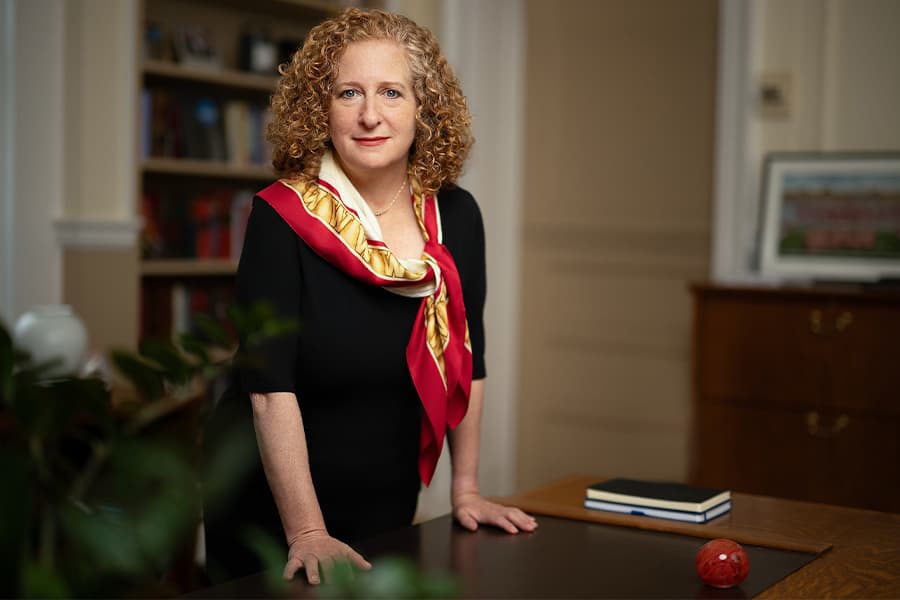 A woman wearing a black dress and a red, gold and white scarf stands at a desk, looking into the camera. She has red, curly hair and red lipstick.