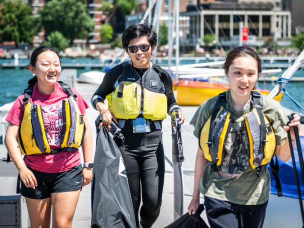 Three people wearing life jackets and carrying sailing gear walk down a pier and get ready to board a sailboat.