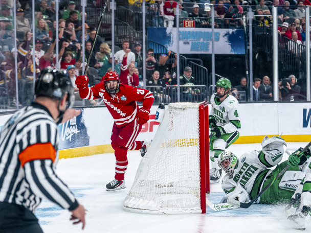 Wisconsin player 21 celebrates his goal with his stick overhead as the North Dakota goalie falls to the ice.