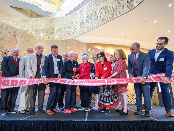 A group of people stand on stage. The Morgridges hold a large pair of red scissors in their hands as they cut a red ribbon that is displayed in front of the large group.