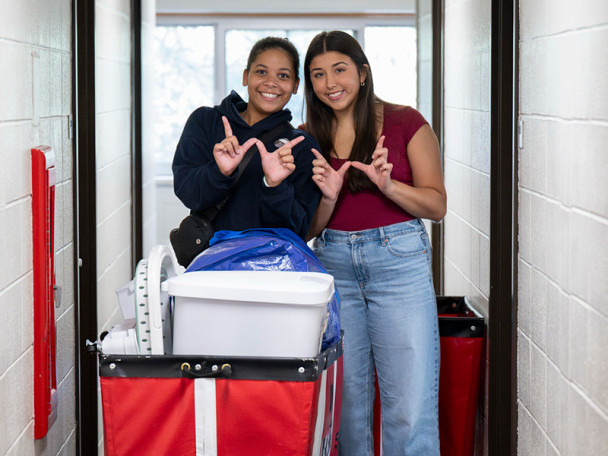 Roommates Leena Brown and Kaylee Katra make Ws with their hands, standing behind a moving cart in the hallway of a residence hall.