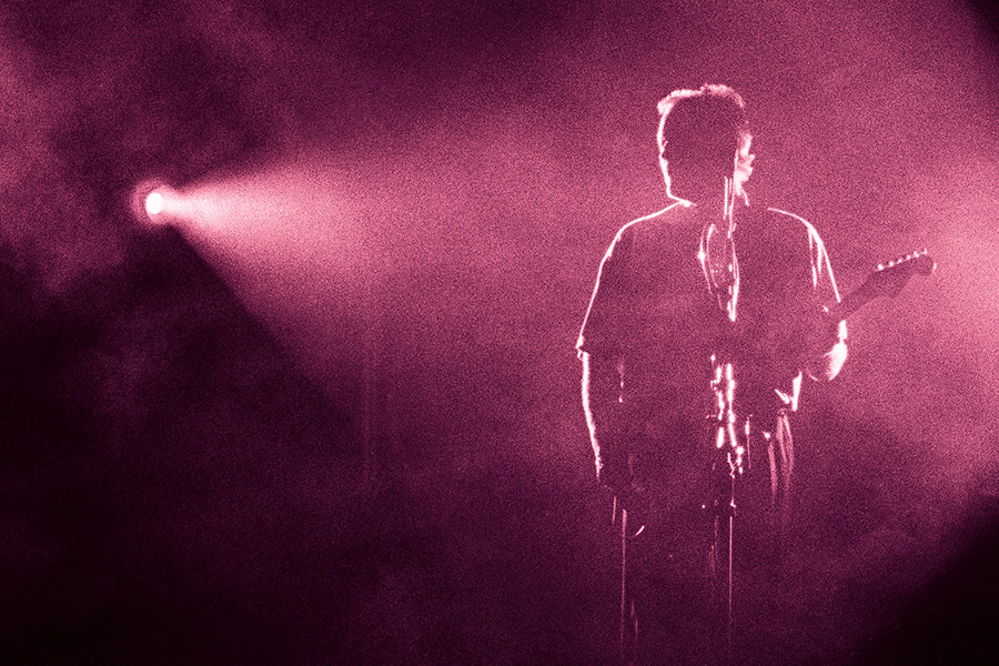 A guitar player, mostly silhouetted, stands on a dark stage with a pink spotlight shining on him.