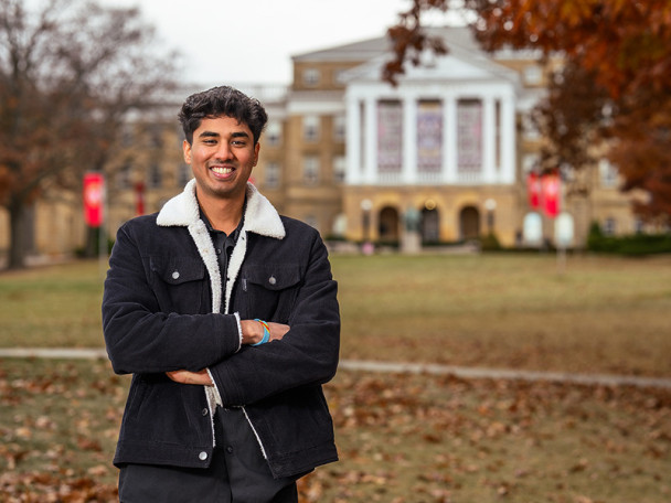 A student wearing a fall coat smiles and stands with his arms crossed. He's standing on Bascom Hill on a late-autumn day.