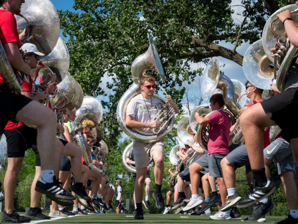 Members of the UW Marching Band tuba section practice marching with high-knee strides outdoors on an athletic field.