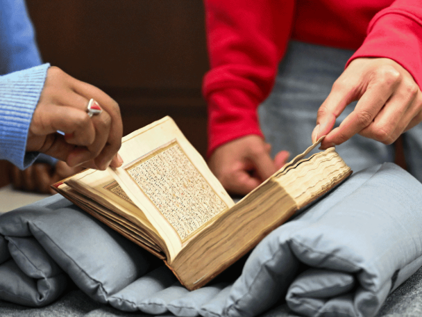 Two students page through an old book that rests on a cushion and table.