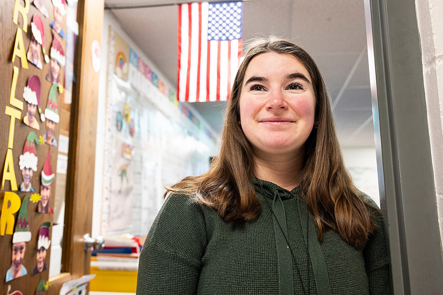 Mackenzie Straub standing in the entrance to her classroom, smiling. The classroom door is decorated with colorful alphabet letters, and an American flag hangs above in the background.